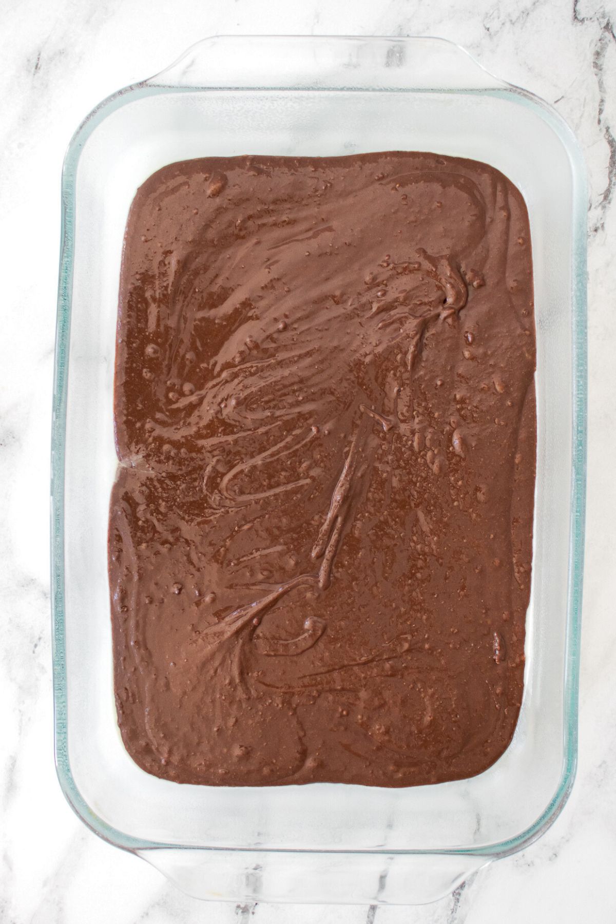 Chocolate brownie batter spread evenly in a clear glass baking dish. 