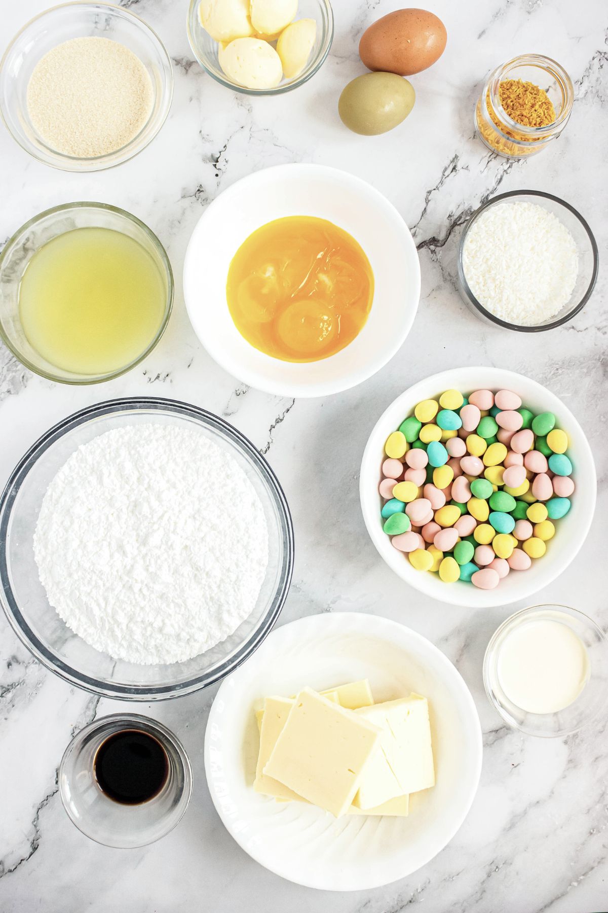 Ingredients needed to make vanilla cupcakes and lemon curd, measured in small glass bowls on a white marble table. 