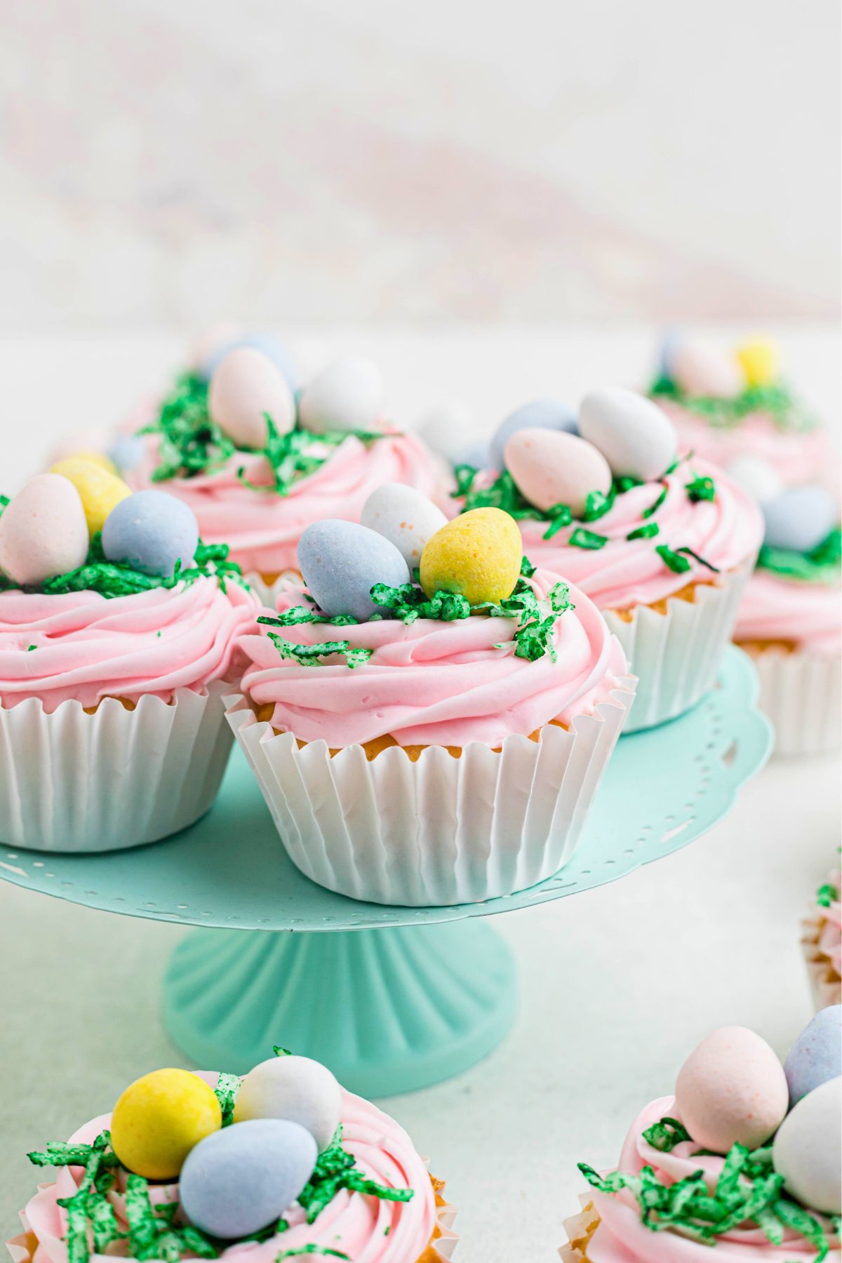 Cute vanilla cupcakes topped with pink frosting, green coconut, and chocolate candy eggs on a light green stand. 