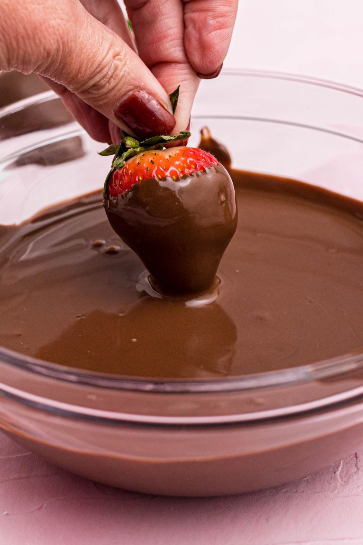 Fresh strawberry being dipped into melted milk chocolate in a clear glass bowl. 