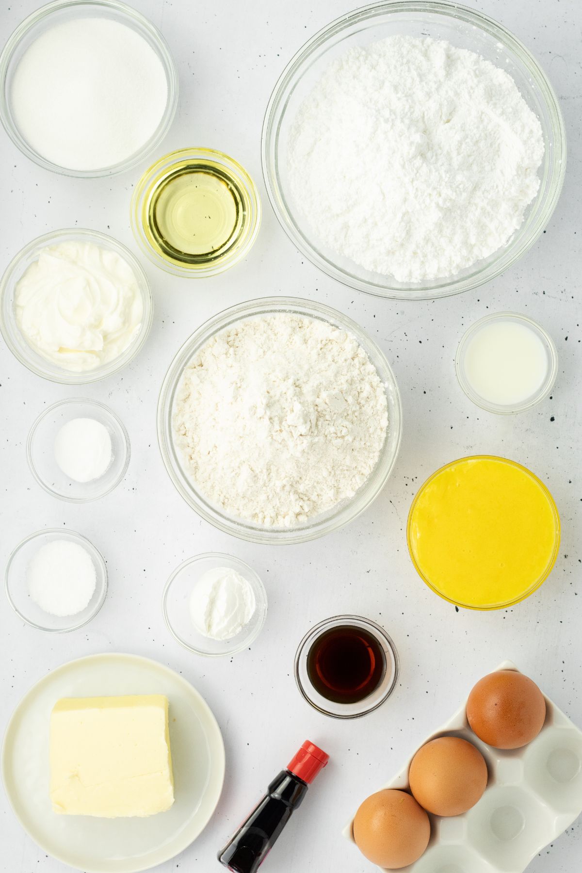 Ingredients needed to make vanilla cupcakes measured into small glass bowls and laid out on a white marble table. 