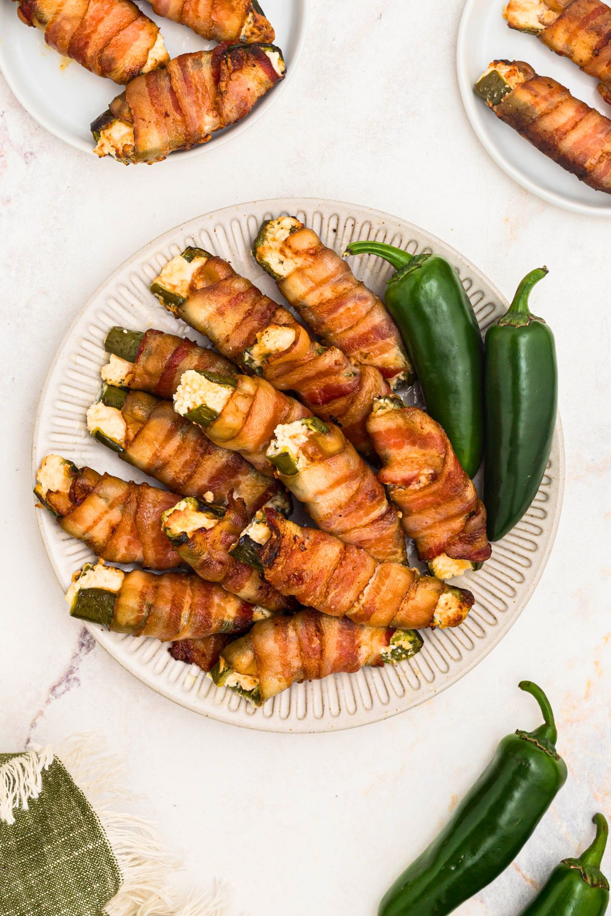 bacon wrapped jalapeno poppers stacked on a white plate with whole peppers next to them. 