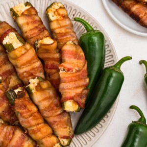 Crispy bacon wrapped jalapenos filled with cheese, stacked on a white plate.