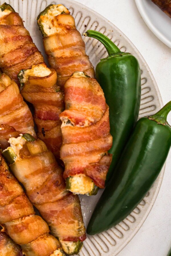 Crispy bacon wrapped jalapenos filled with cheese, stacked on a white plate.