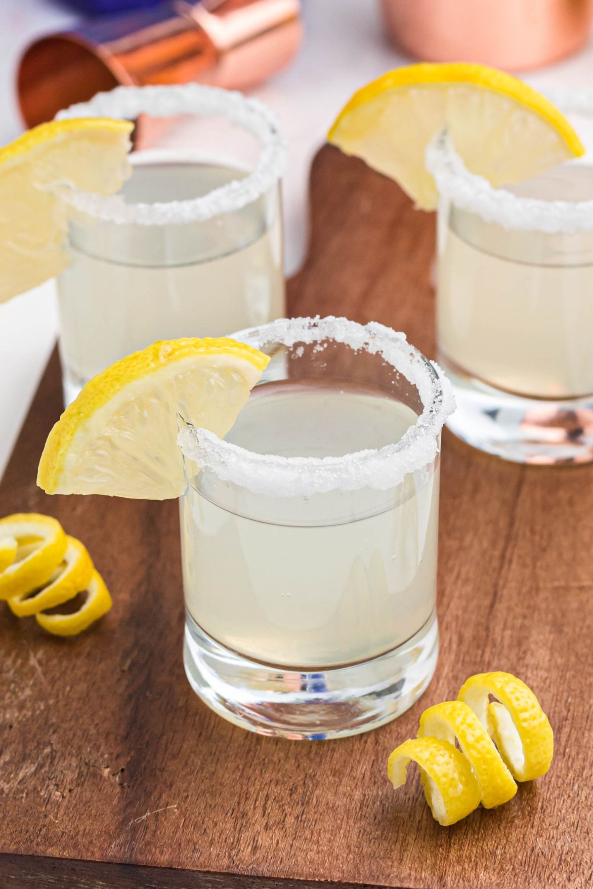 Sugar rimmed shot glass with lemon wedge for garnish, and lemon curls in front of glasses, after being filled with lemon drop shots. 