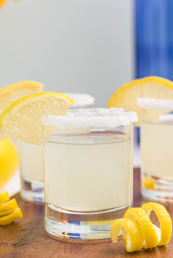 Sugar rimmed shot glass with lemon wedge for garnish, and lemon curls in front of glasses, after being filled with lemon drop shots.