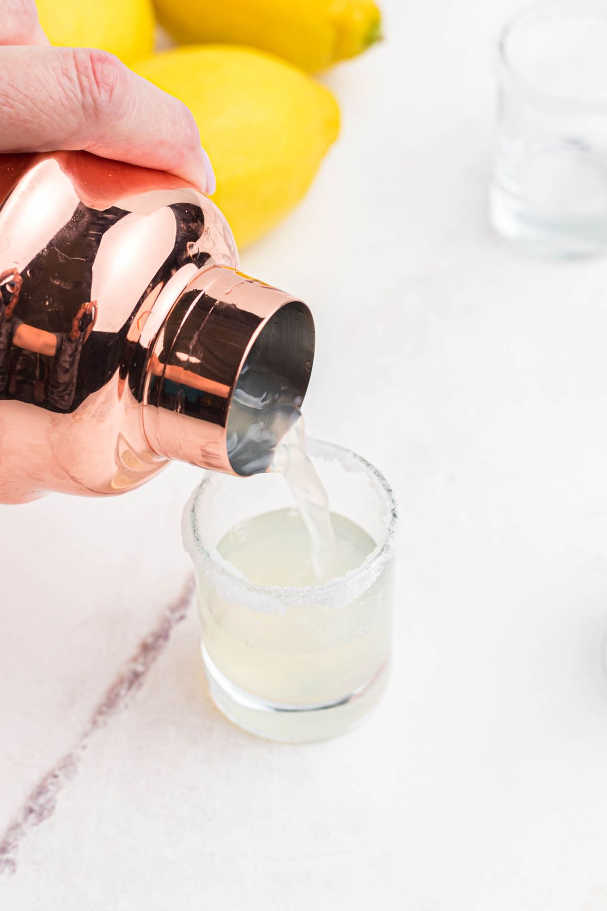 Vodka and lemon juice with simple syrup being poured from a cocktail shaker into a sugar rimmed shot glass.