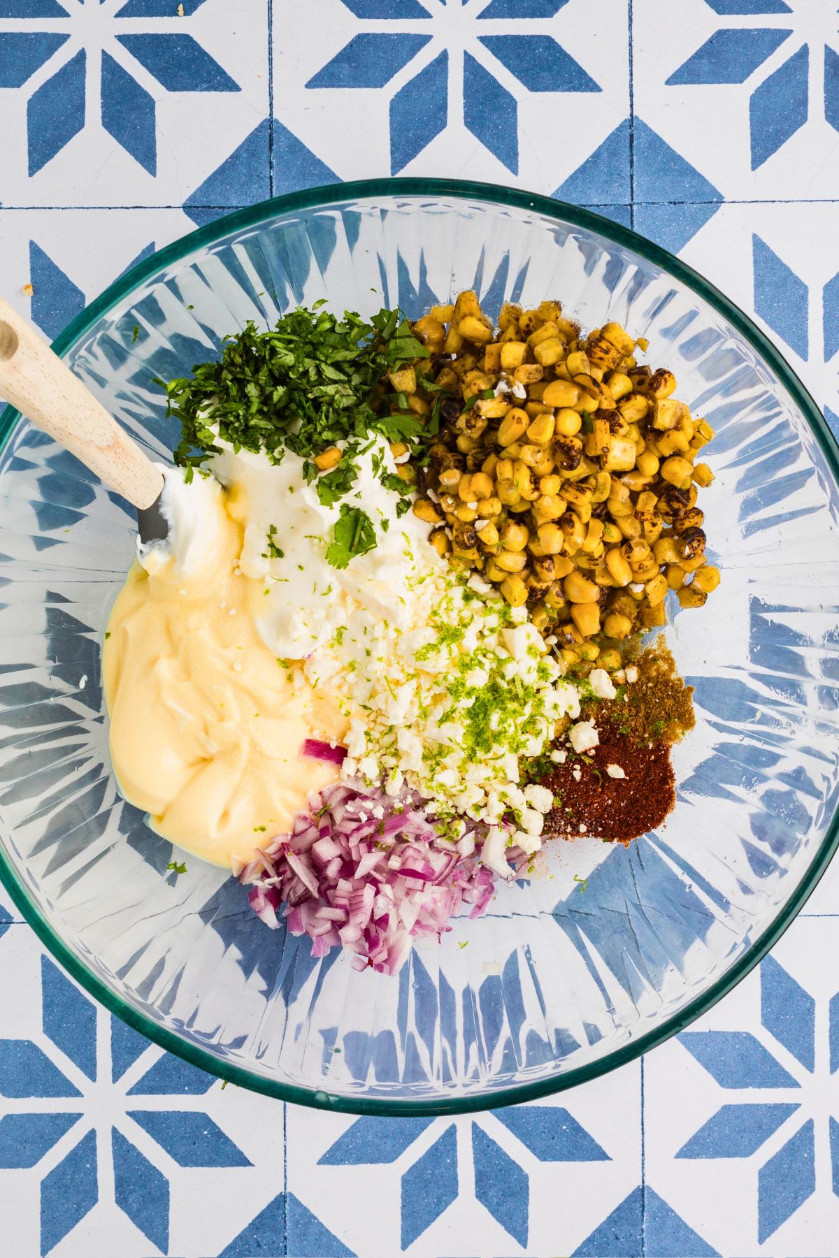 Spices, cheese, roasted corn, and other ingredients mixed together in a clear  mixing bowl, to make a corn dip. 