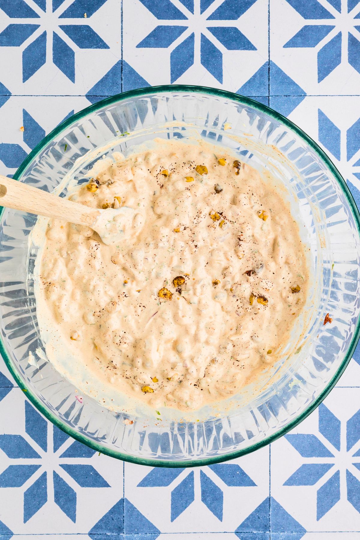 Corn dip ingredients mixed together in a clear glass bowl on a blue tiled table. 