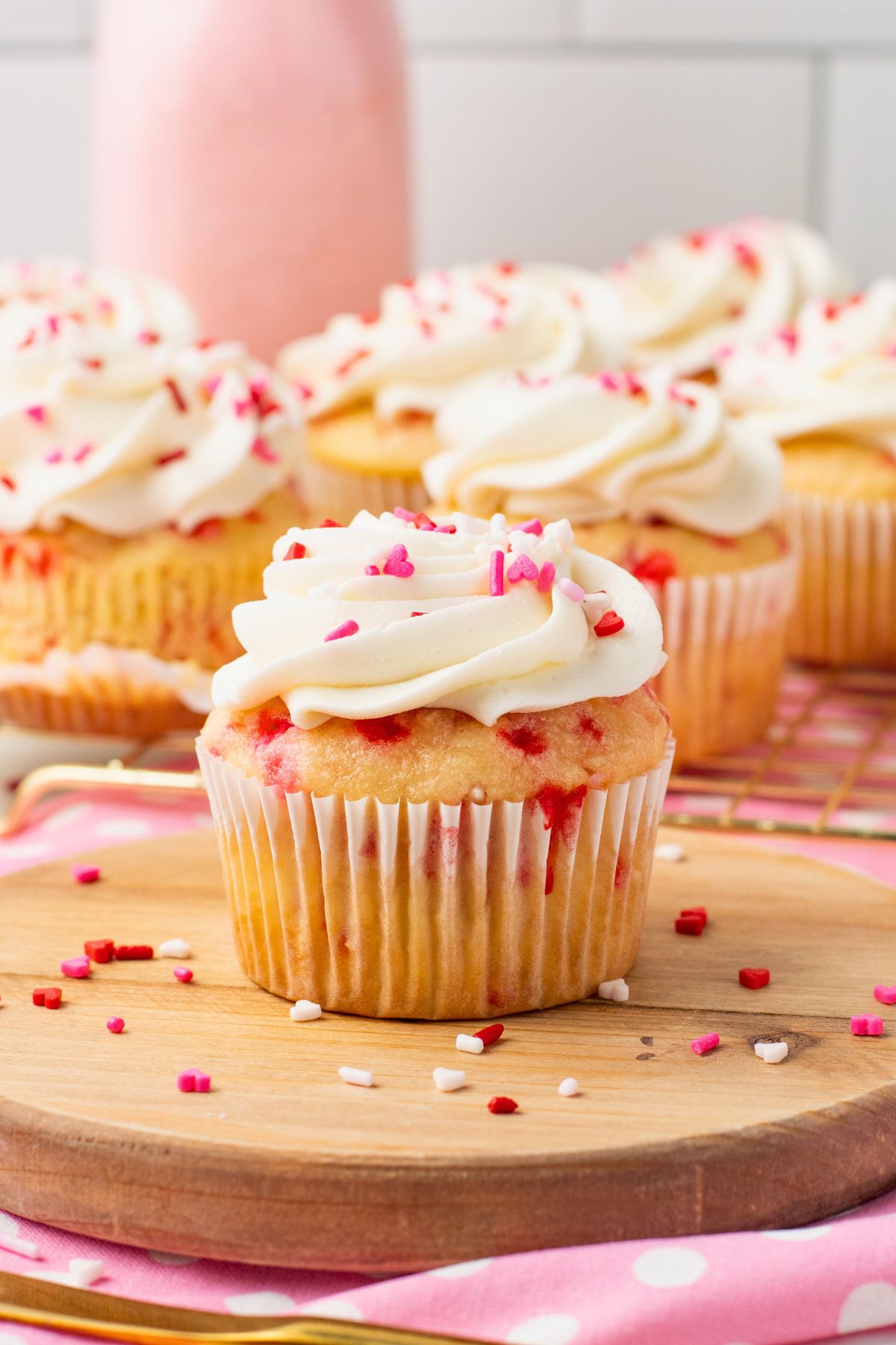 Vanilla cupcakes filled with pink sprinkles topped with white frosting and pink sprinkles with pink milk in the background. 