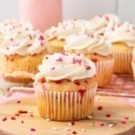White cupcakes with pink sprinkles baked in the batter, then frosted with white frosting and topped with pink sprinkles.