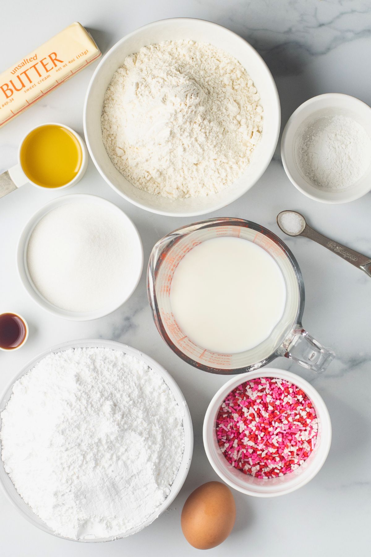 Ingredients needed to make vanilla cupcakes from scratch with pink sprinkles in small white bowls. 