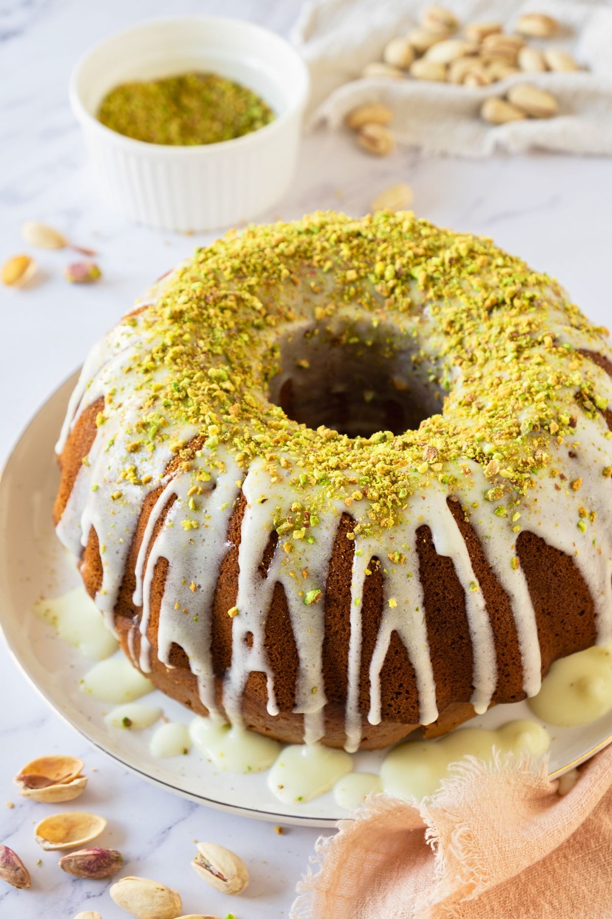 Pistachio Bundt cake on a plate with a bowl of chopped pistachios behind it on a marble table. 