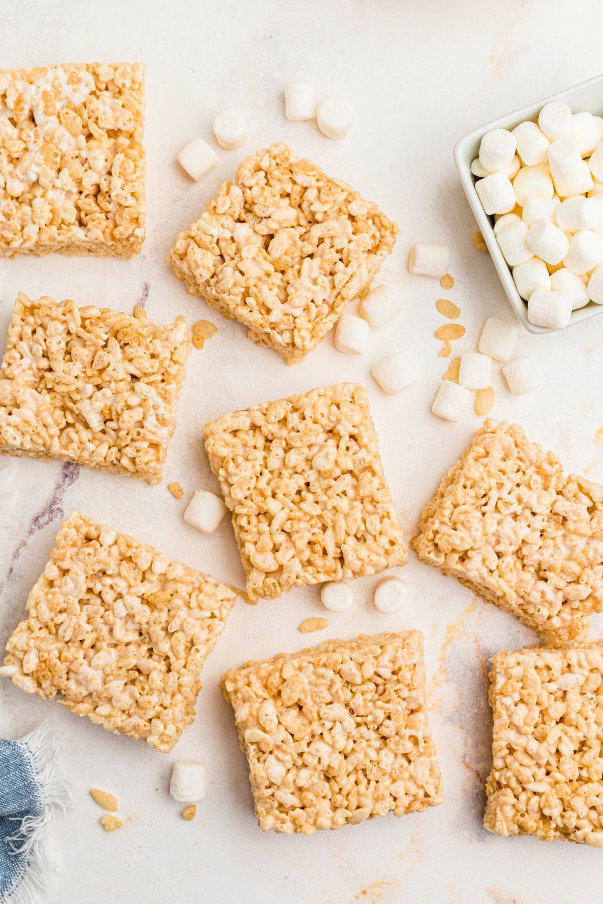 Rice krispie treats cut into squares on a marble table with mini marshmallows in a small bowl. 