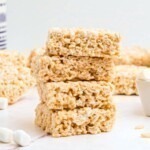 Four rice krispie treats stacked together on a white marble table with mini marshmallows on the table.