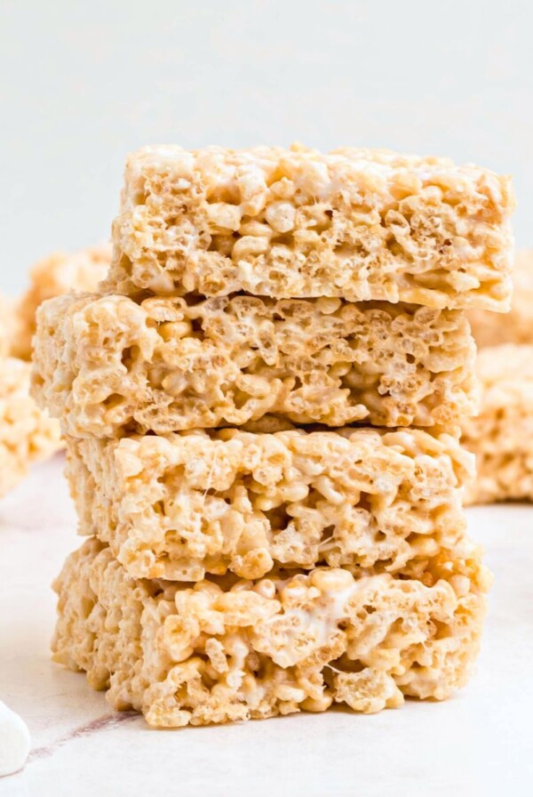 Four rice krispie treats stacked together on a white marble table with mini marshmallows on the table.