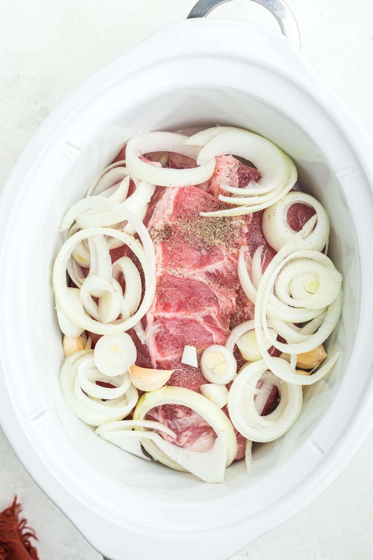 Uncooked ribs in a white slow cooker, topped with onion slices, garlic, and seasonings. 