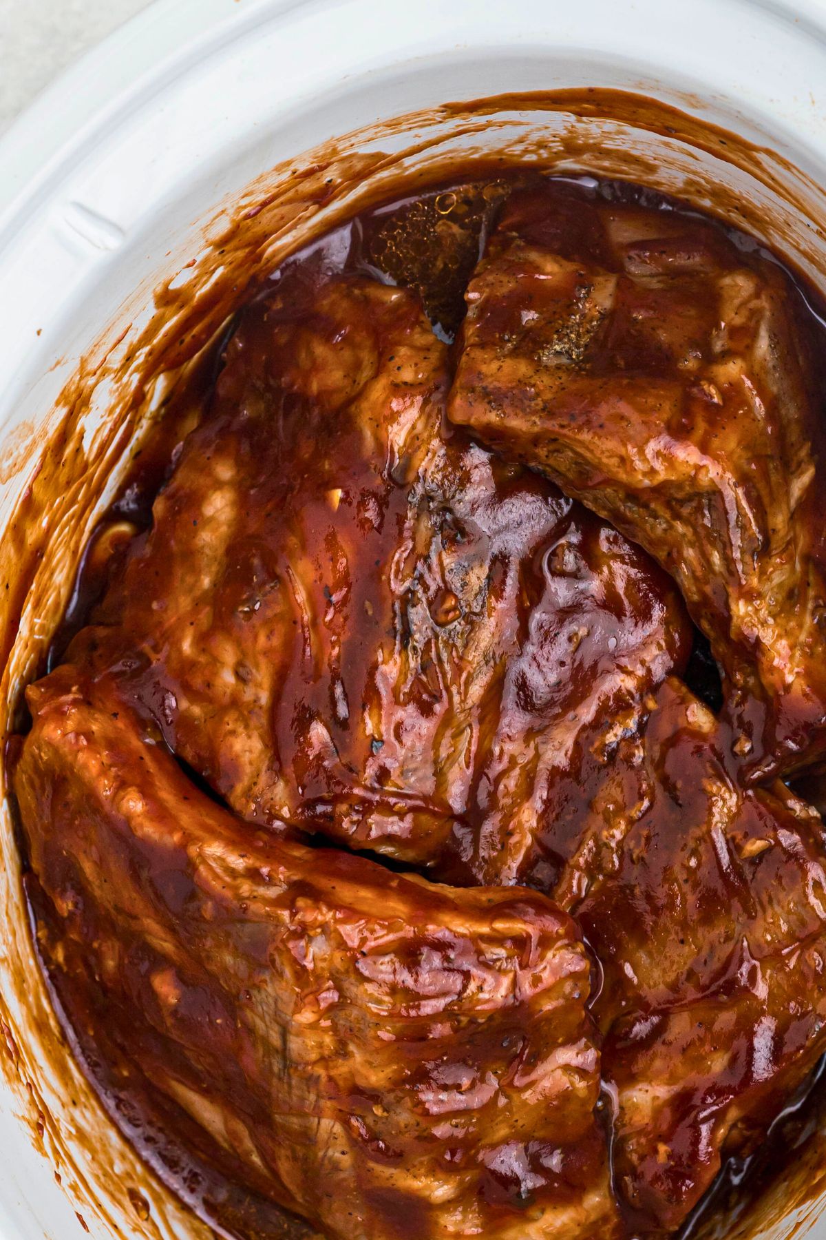 Juicy baby back ribs in a white slow cooker, tossed with BBQ sauce. 