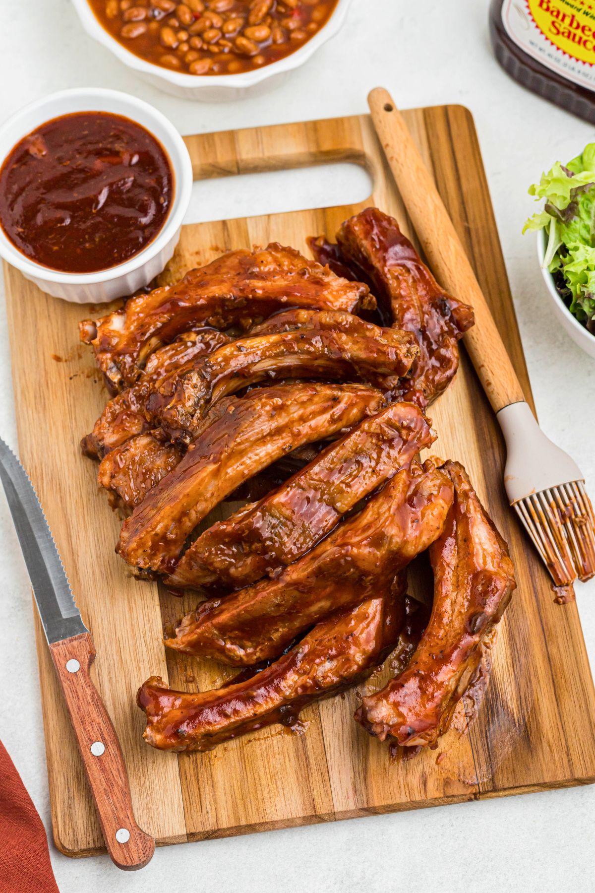 Juicy ribs cut individually, on a wooden cutting board with a steak knife and brush next to them. 