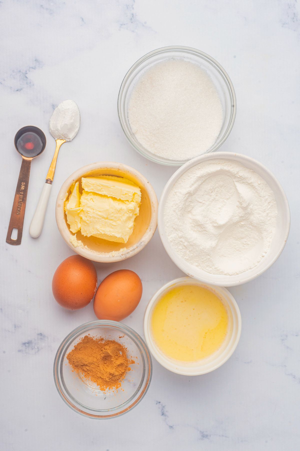 Ingredients needed to make cupcakes measured in small bowls on a white marble table. 