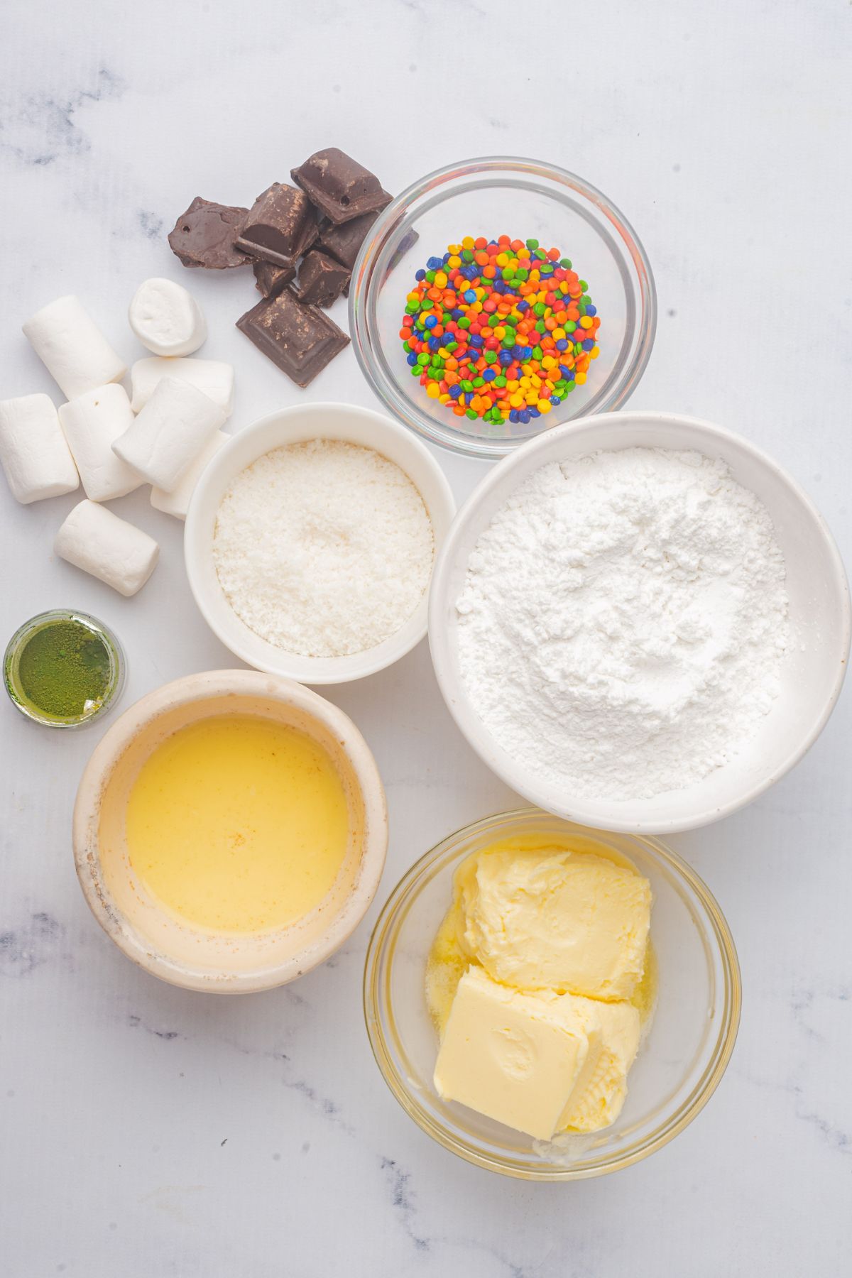 Frosting ingredients and decorations measured into small glass bowls on a white marble table. 