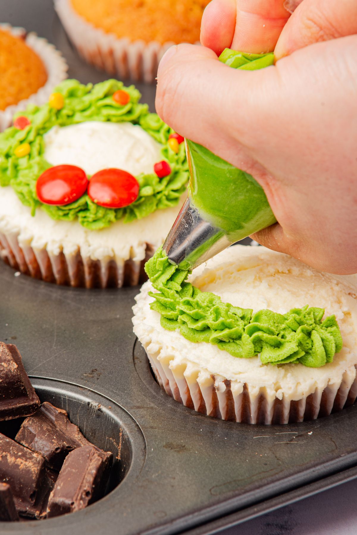 Wreath cupcakes being decorated with green frosting and red candies. 