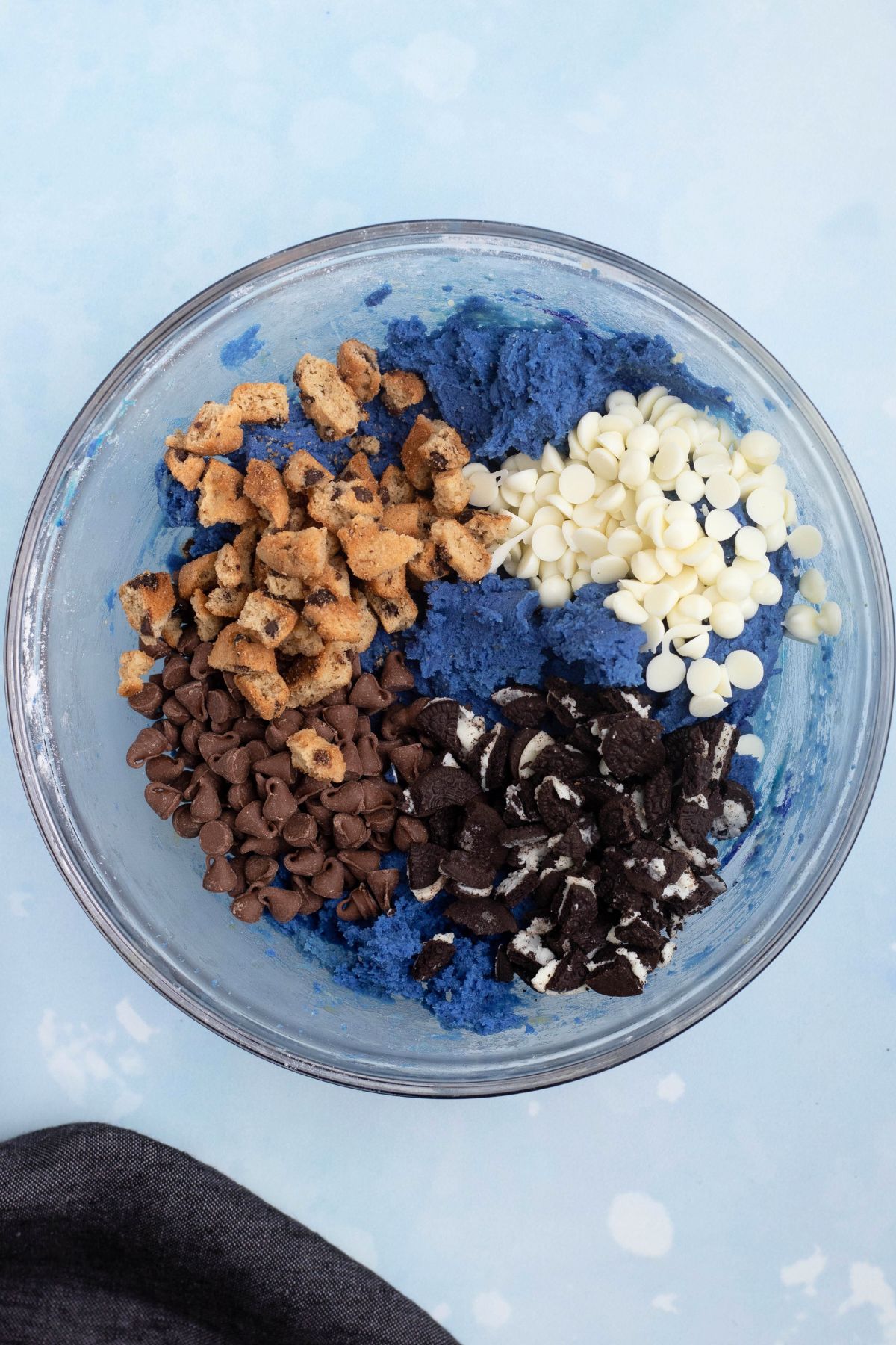 Chopped cookies and chips being mixed in with cookie dough in a clear glass bowl.