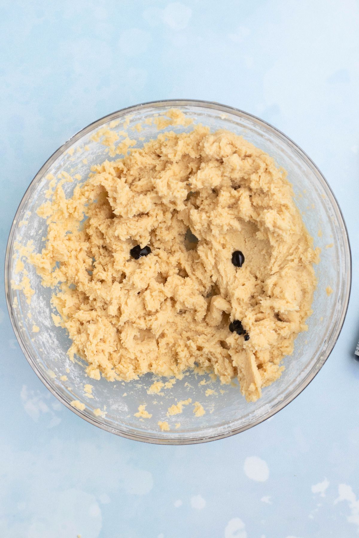 Cookie dough being mixed with royal blue and violet food colors in a clear glass mixing bowl.