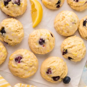 Lemon blueberry cookies on parchment paper with lemon wedges and blueberries scattered next to them.