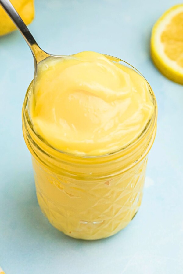 Bright yellow lemon curd in a small jar with a spoon being scooped out.