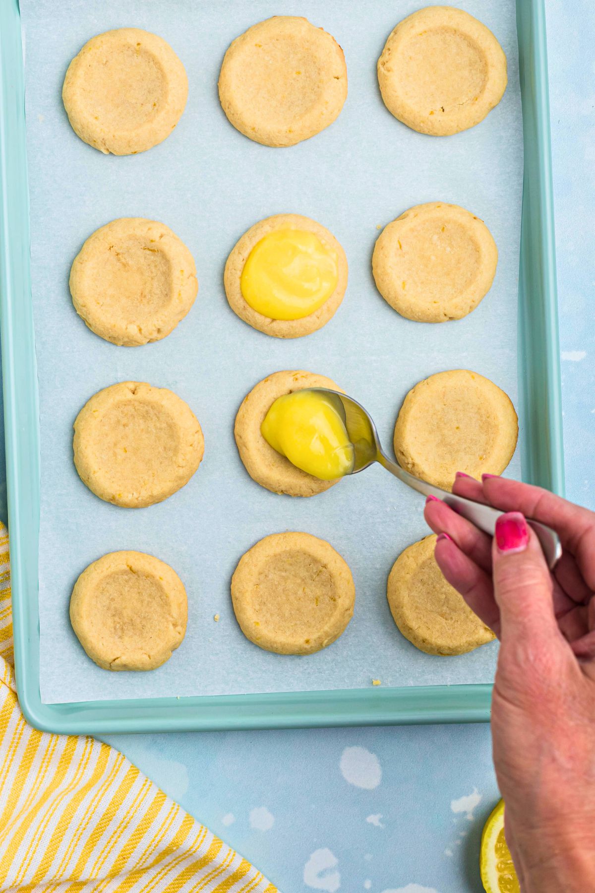 Shortbread cookies being topped with a spoonful of lemon curd, on a baking sheet. 