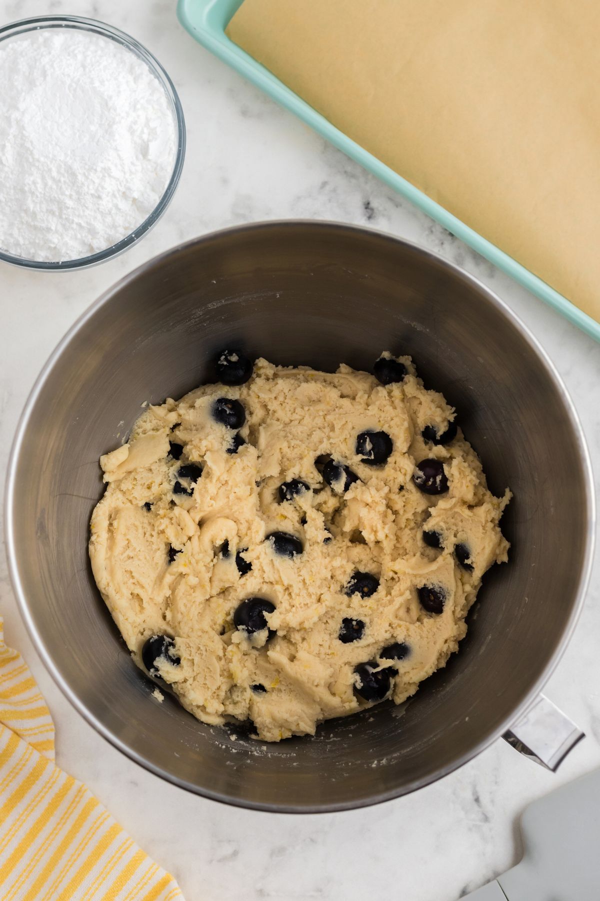 Cookie dough combined with blueberries in a mixing bowl, next to a baking sheet lined with parchment paper. 