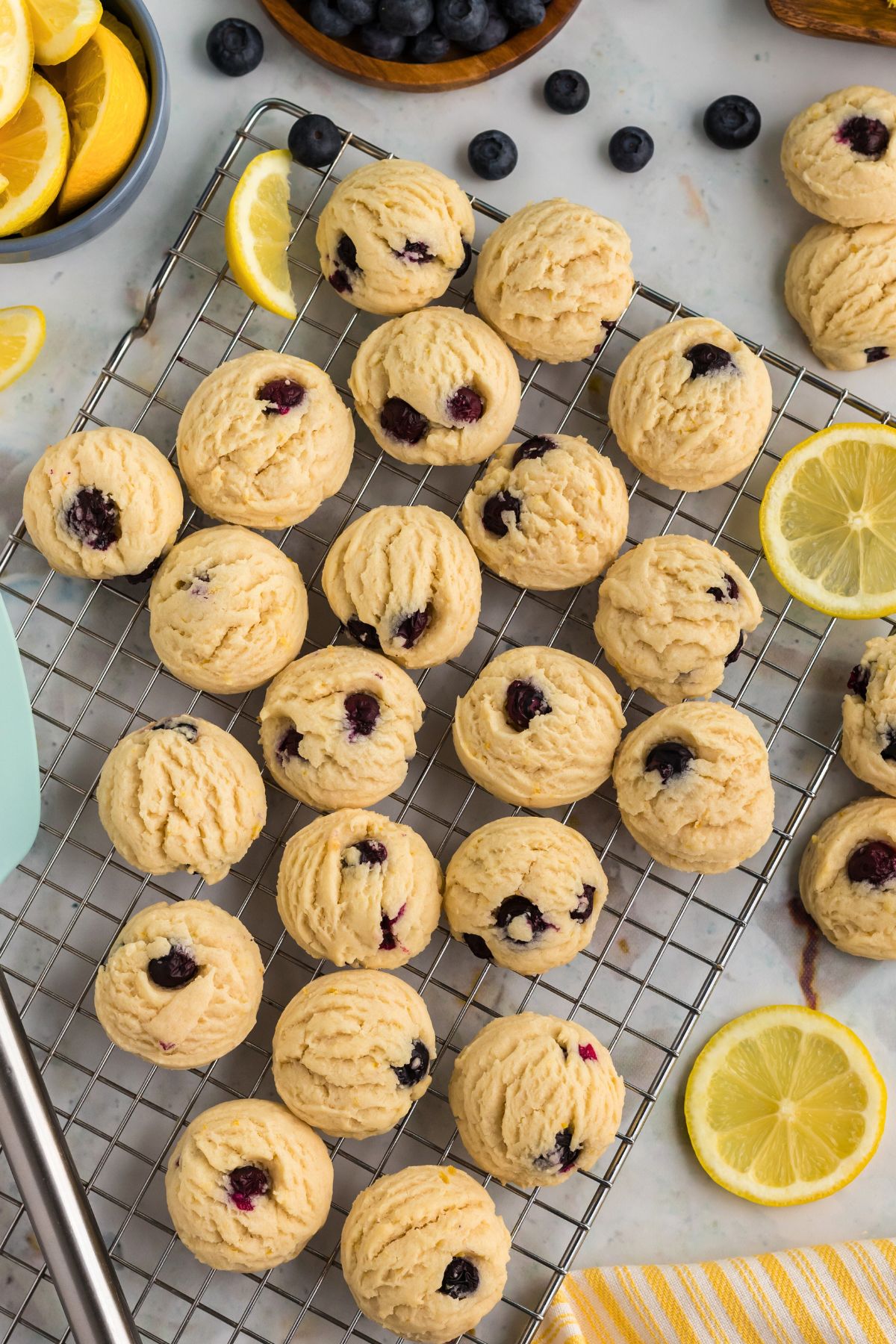 Lemon blueberry cookies on a wire rack with blueberries and lemon wedges on the table. 