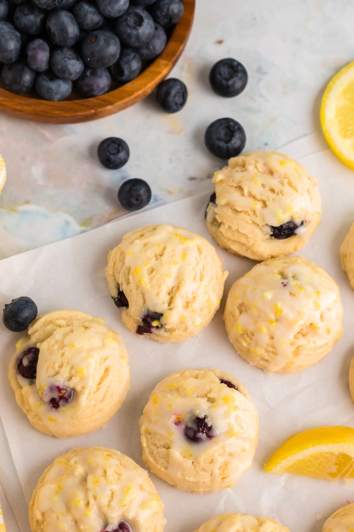 Lemon blueberry cookies on parchment paper with lemon wedges and blueberries scattered next to them. 