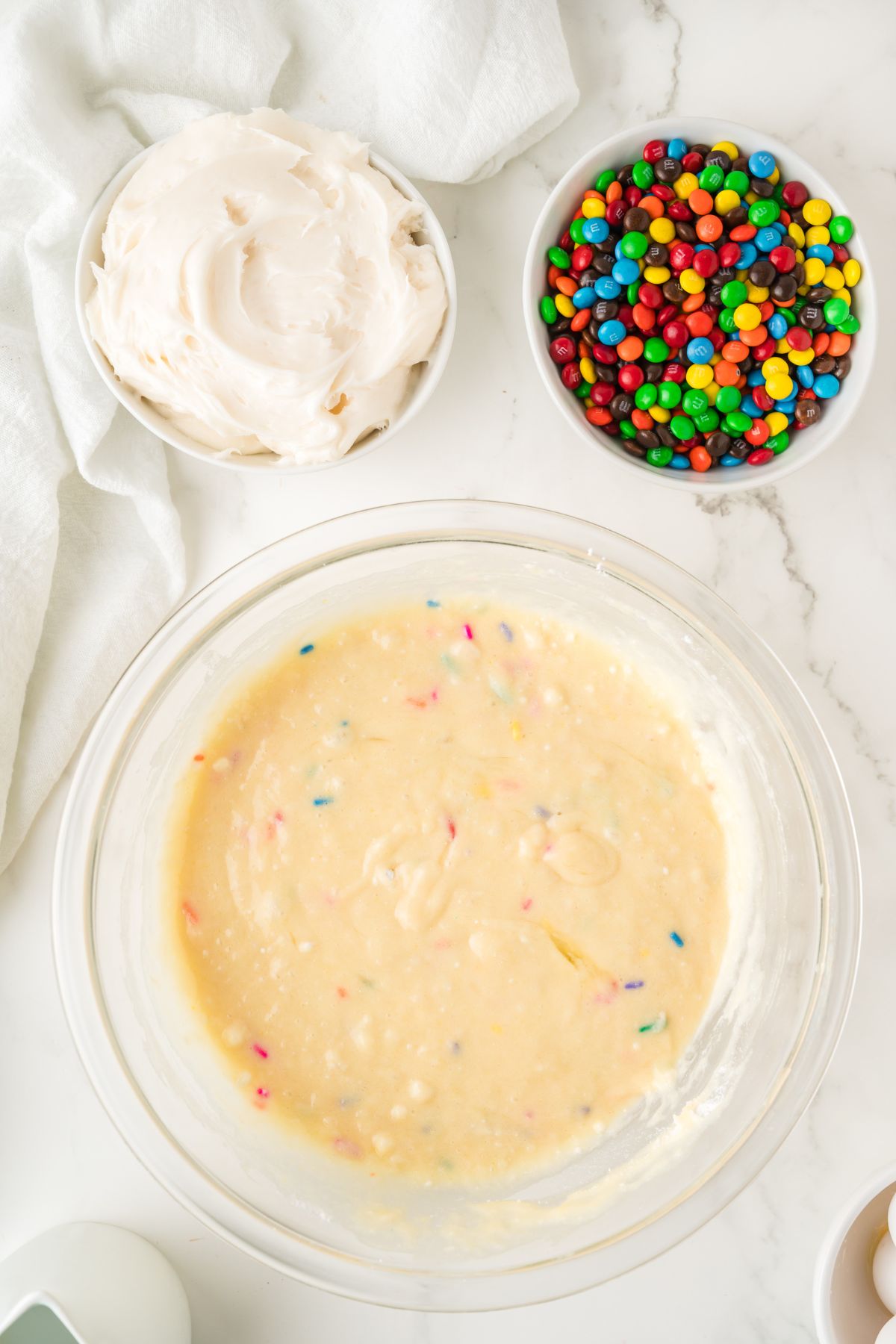 Cake batter mixed in a large glass bowl with frosting and m and m's on a marble. 