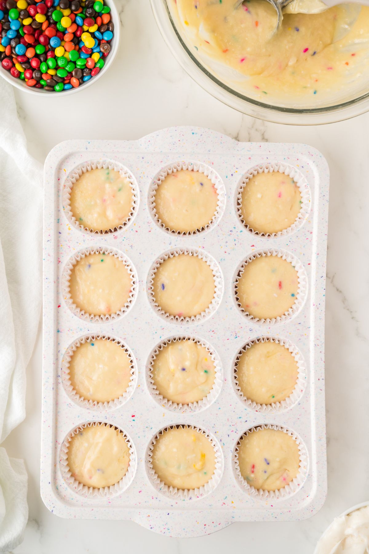 Cupcake batter poured into muffin pan with mini m and m's in a bowl nearby. 