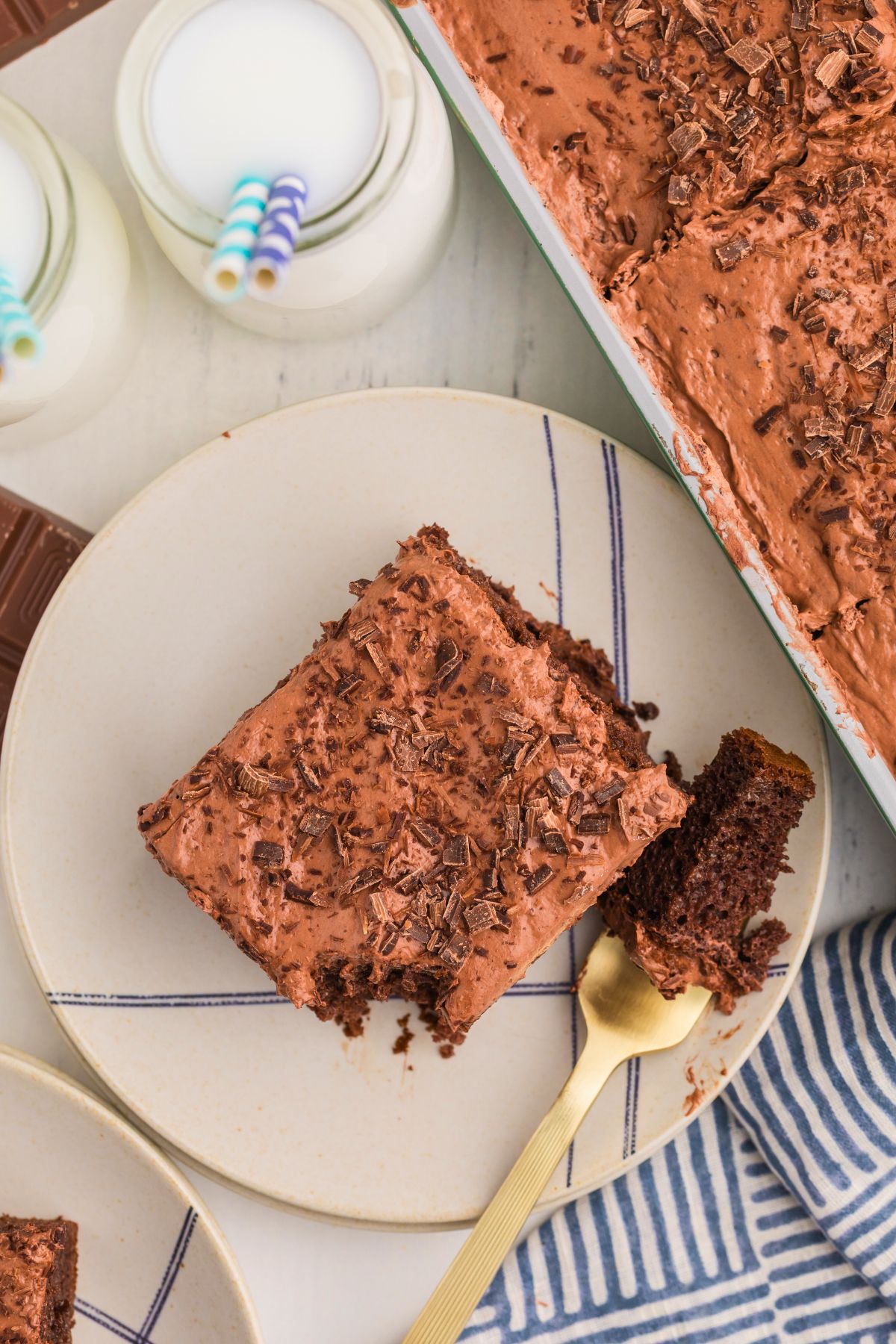 Slice of chocolate poke cake on a white plate in front of glasses of milk with blue straws.