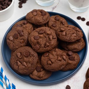 Double chocolate chip cookies stacked on a blue plate with chips and milk on the table.