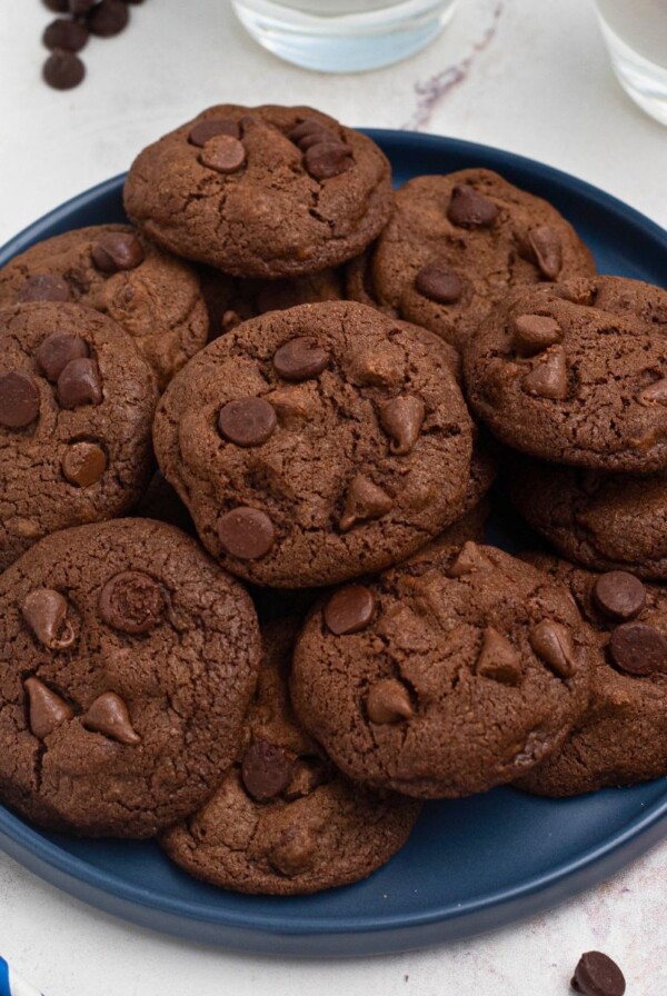 Double chocolate chip cookies stacked on a blue plate with chips and milk on the table.