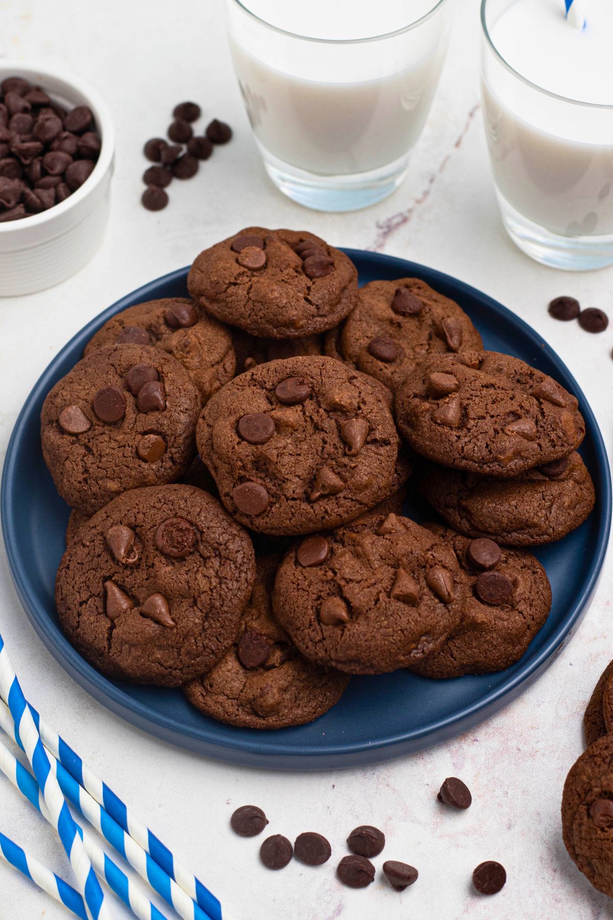 Double chocolate chip cookies stacked on a blue plate with chips and milk on the table. 