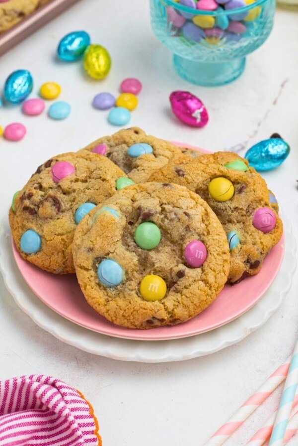 Golden chocolate chip cookies with pastel candies on plates, in front of candy dish and glasses of milk.