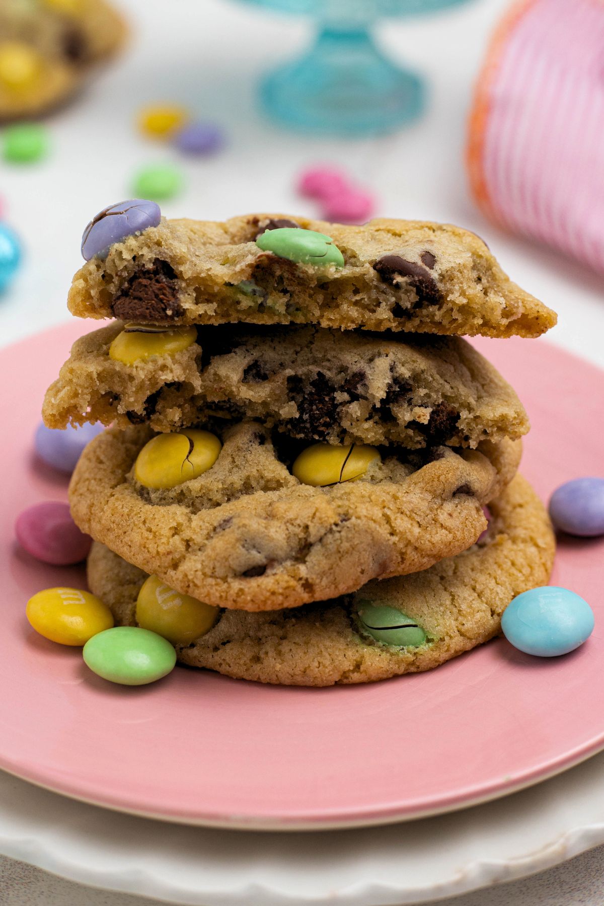 Stacked chocolate chip cookies with pastel m&m's on a pink plate with the top cookie broken in half.