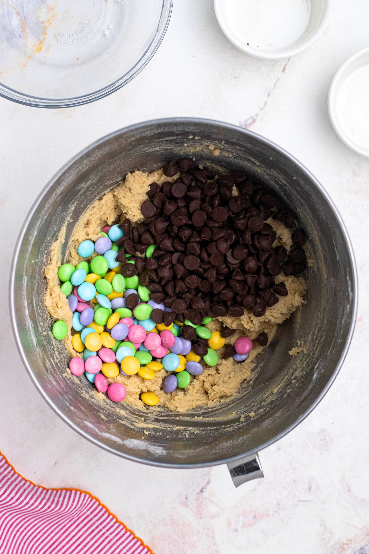 Chocolate chips, pastel m&m's and cookie dough being mixed together in a silver mixing bowl.