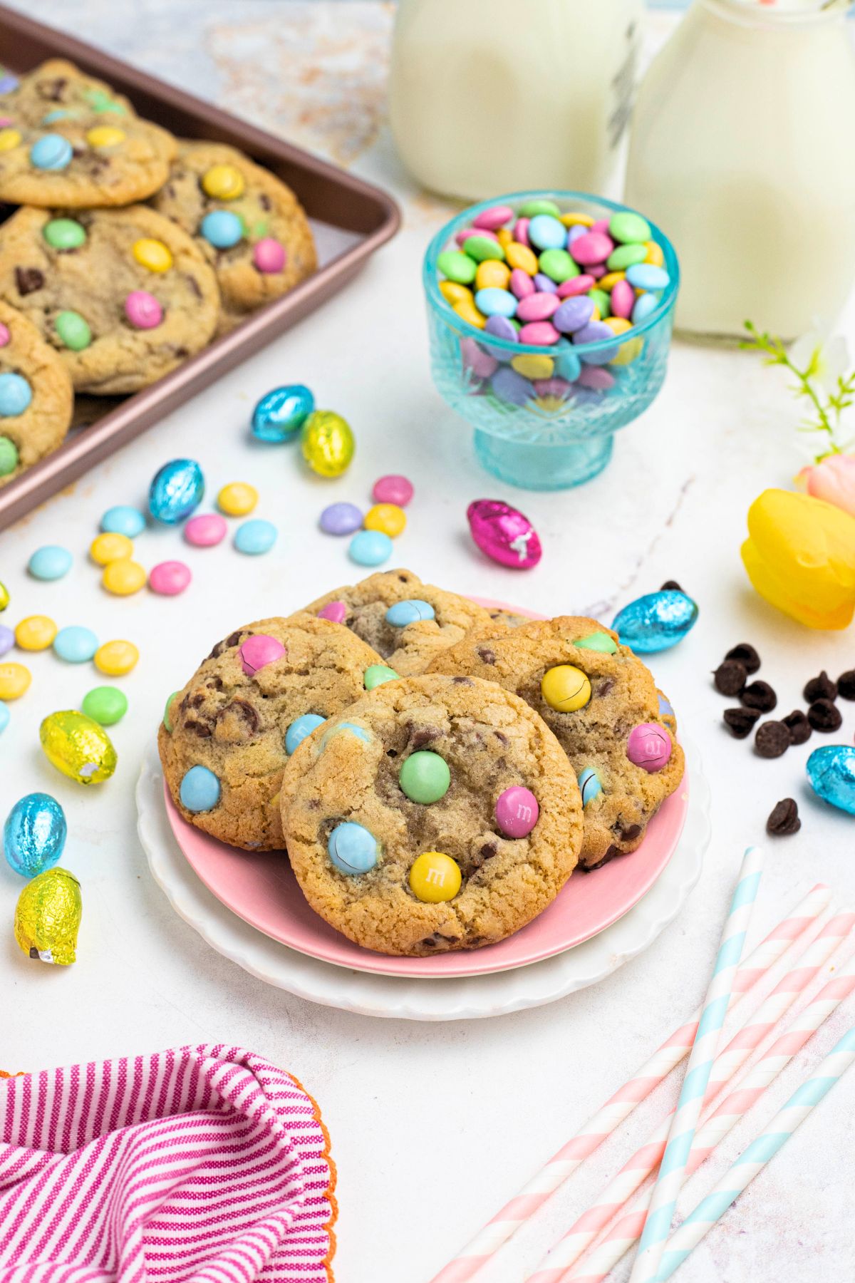 Golden chocolate chip cookies with pastel candies on plates, in front of candy dish and glasses of milk.