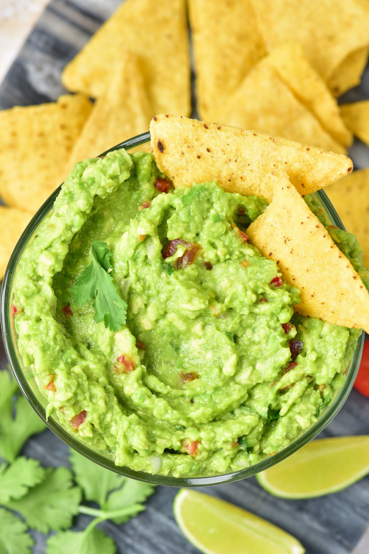 Green creamy guacamole in a clear glass bowl with triangle tortilla chips and lime wedges around the bowl. 