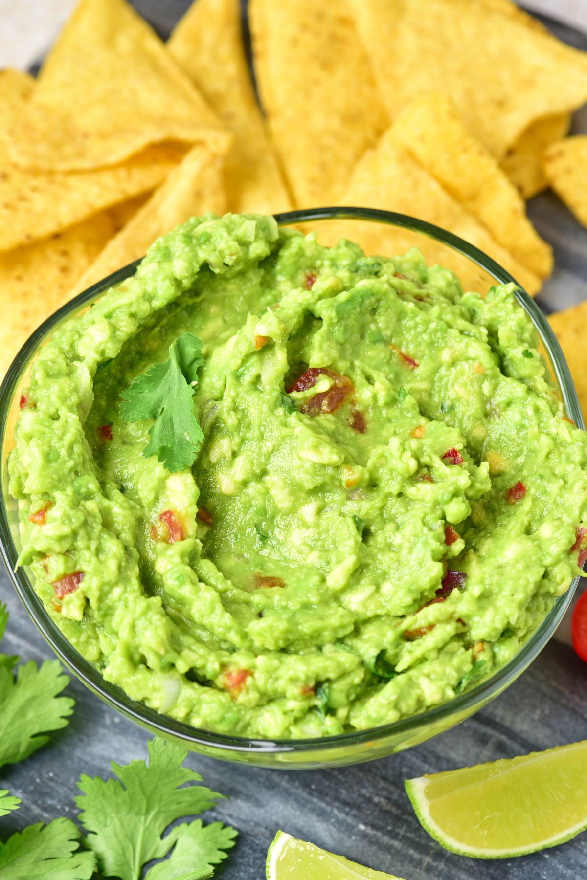 Stirred guacamole in a clear glass bowl on a cutting bowl, with lime wedges and tortilla chips scattered around. 