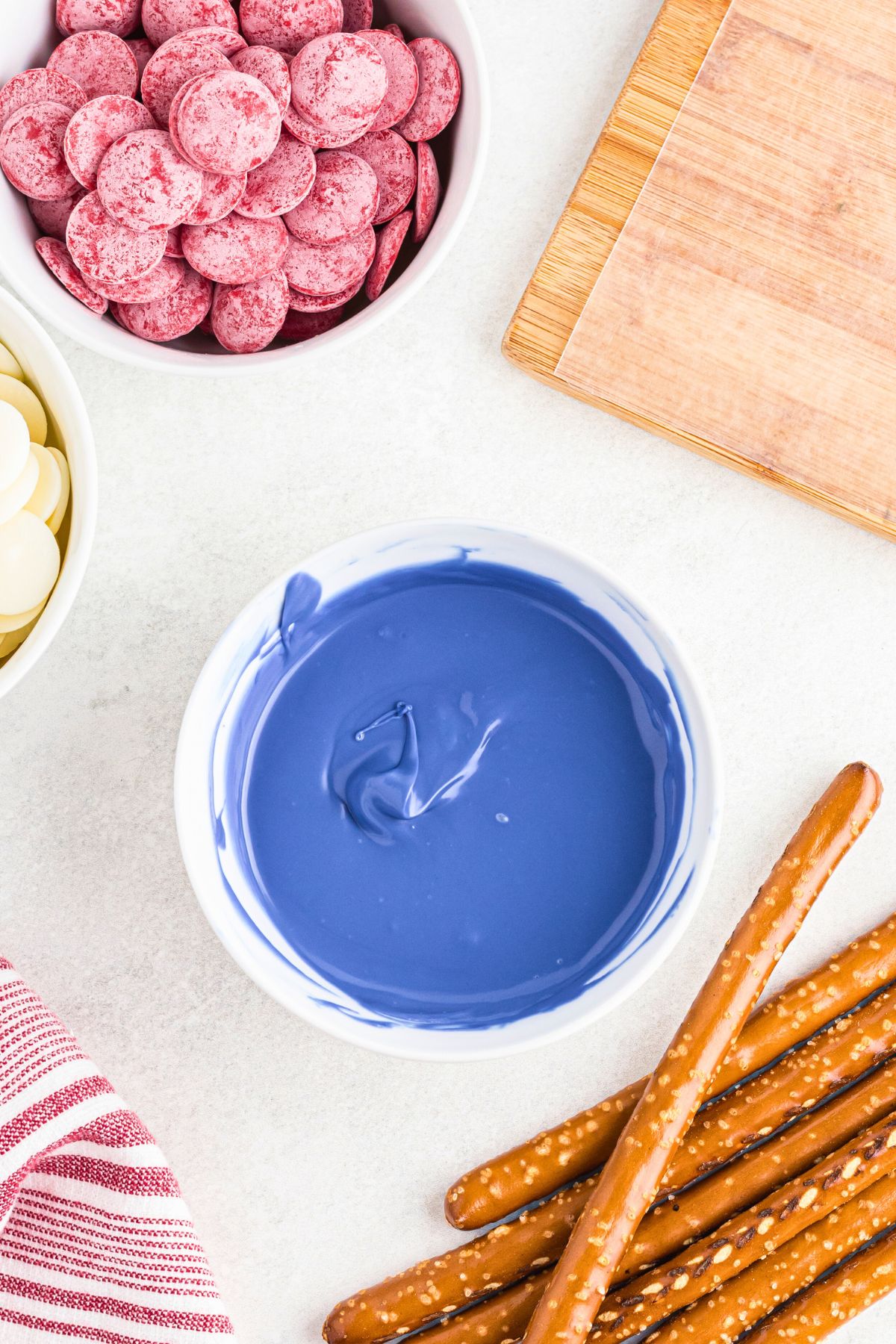 Bright blue melting chocolate melted in a small white bowl in front of pretzel rods, before being dipped. 