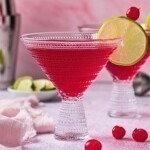 Red pomegranate martini in a clear cocktail glass with a lime wedge and cherry for a garnish.