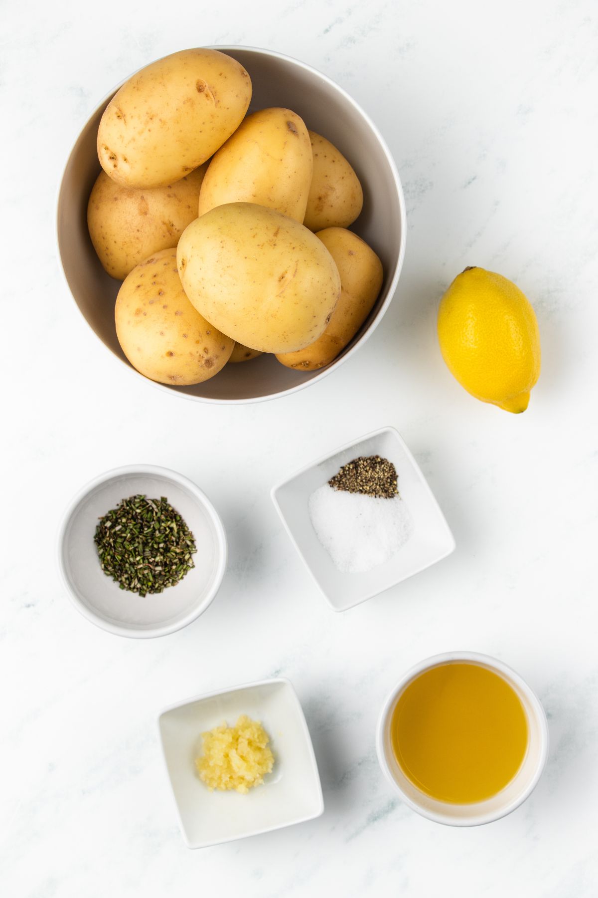 Ingredients needed to make roasted potatoes measured in small bowls on a white marble table. 
