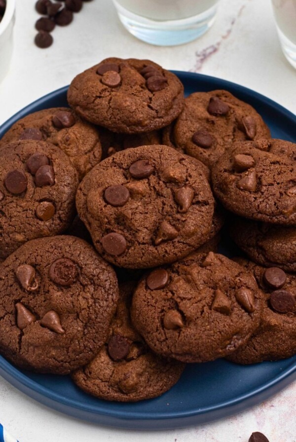 Double chocolate chip cookies stacked on a blue plate with chips and milk on the table.